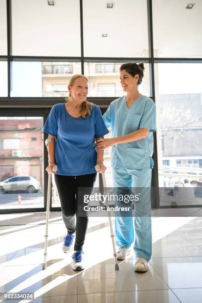 senior woman using crutches coming into the clinic for physiotherapy and therapist helping her - crutch stock pictures, royalty-free photos & images