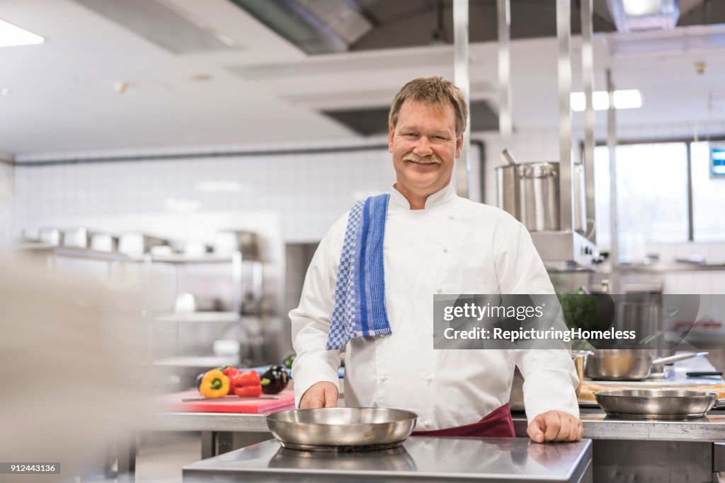 Ein Koch in seiner Küche hält eine Pfanne und lächelt in die Kamera
