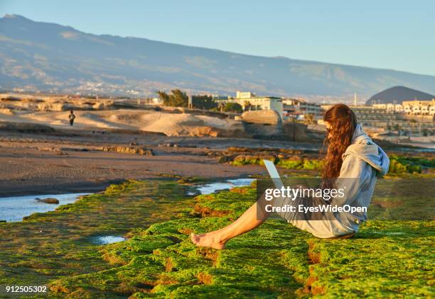 Woman sits alone on the rocks with colorful seaweed working with a notebook at the beach on January 19, 2018 in El Medano, Tenerife, Spain. Model...