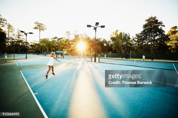 mature woman hitting forehand during early morning tennis match - schlägersport stock-fotos und bilder