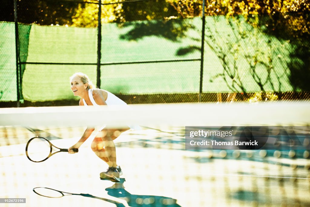 Smiling mature female tennis player preparing to hit return at net during early morning tennis match