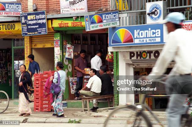 Street scene with signs in English and Nepali advertising film processing, and computer and language classes. Men sit outside a grocery shop, women...