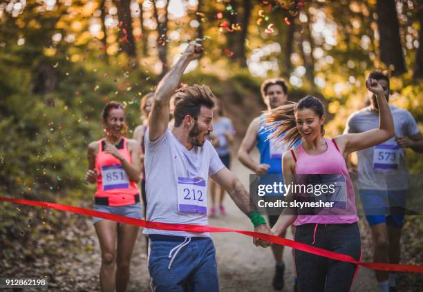 happy athletic couple holding hands while running through finish line together. - finish line stock pictures, royalty-free photos & images