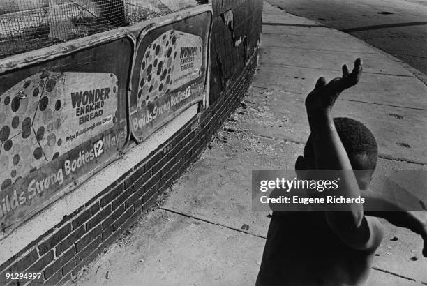 View of young boy as he strikes a karate pose on a sidewalk, Dorchester, Massachusetts, 1975. A low brick wall features a pair of advertisements for...