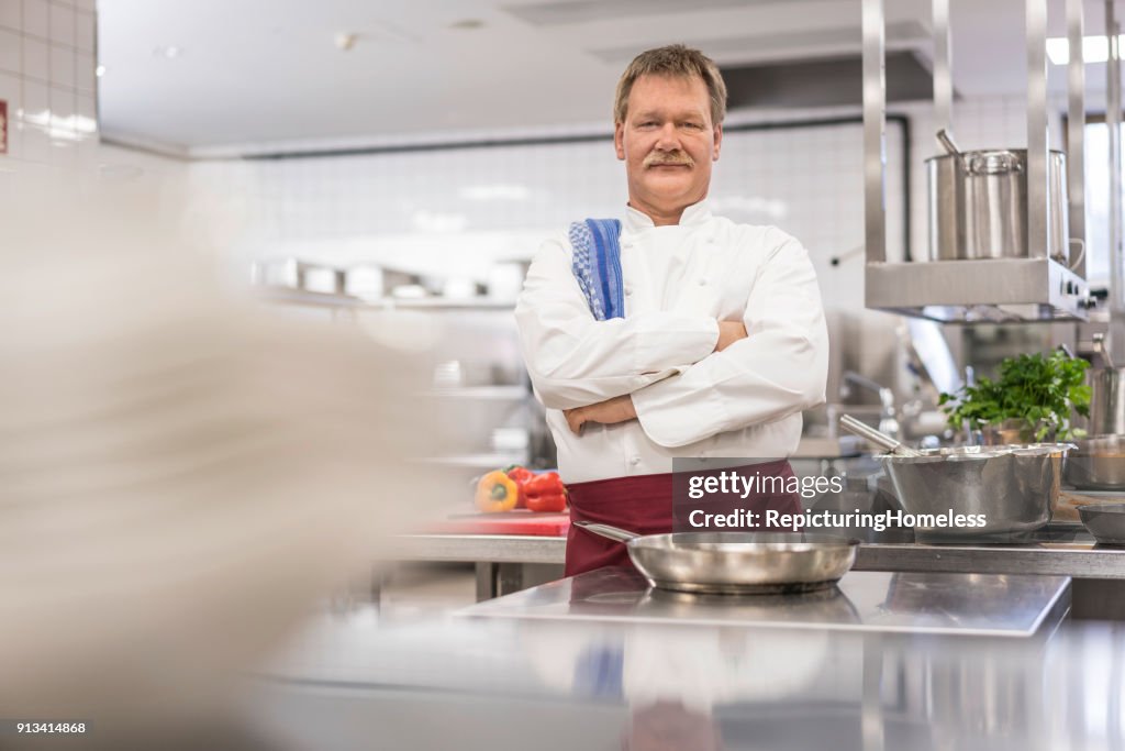 Un chef en la cocina de pie delante de una bandeja con los brazos cruzados y mirando a la cámara