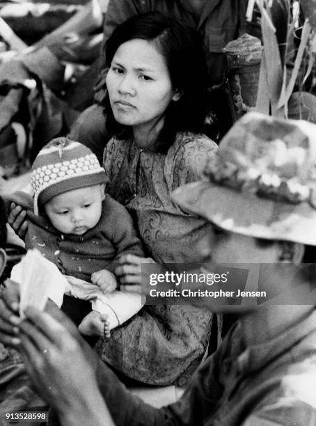 View of an unidentified woman as she holds a child in her lap and watches as an Army of the Republic of Vietnam) soldier plays card at a remote base,...