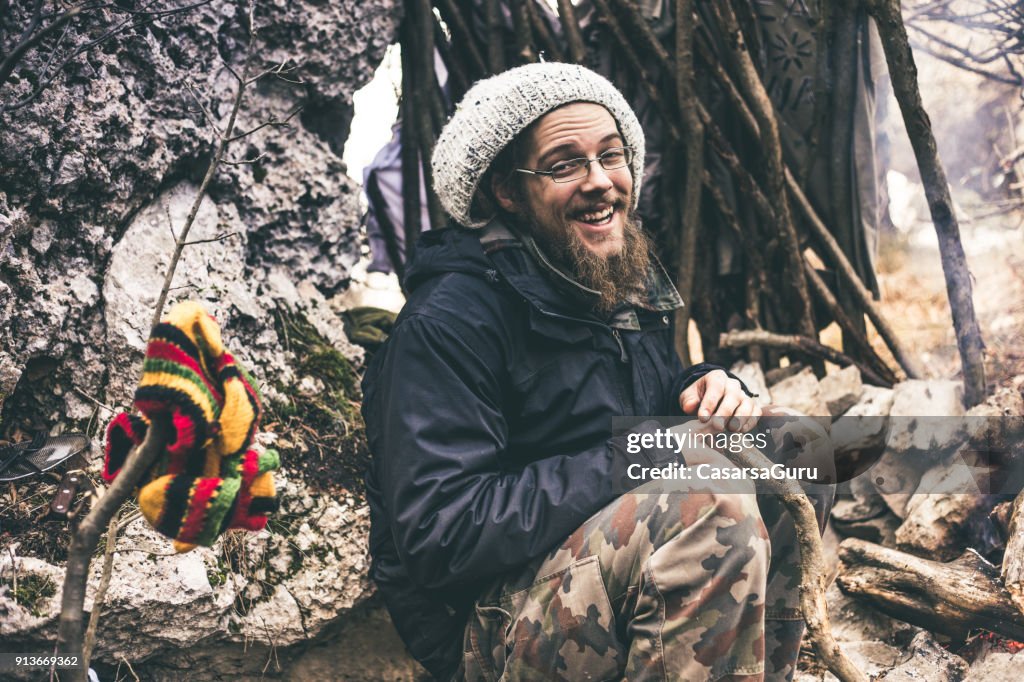 Smiling Young Man with Dreadlocks Sitting By a Fireplace in Nature