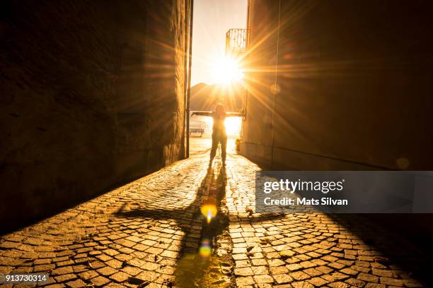 woman with arms outstretched in a narrow street with sunbeam and shadow - narrow stock pictures, royalty-free photos & images
