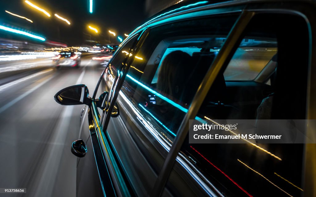Long exposure photograph of a black German car driving on Mallorca, left windows with reflections in foreground, rear view mirror in middle ground and motion blurred background.