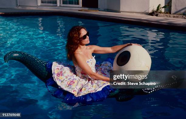 Portrait of American actress Catherine Bach, in sunglasses and a strapless dress, as she sits on an inflatable alligator in a swimming pool, Los...