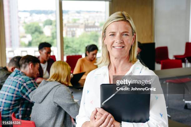 portrait of mature female lecturer holding folder in classroom, smiling - teacher with folder stock pictures, royalty-free photos & images