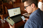 Attractive man in glasses working with multiple electronic internet devices. Freelancer businessman has laptop and smartphone in hands and laptop on table with charts on screen. Multitasking theme