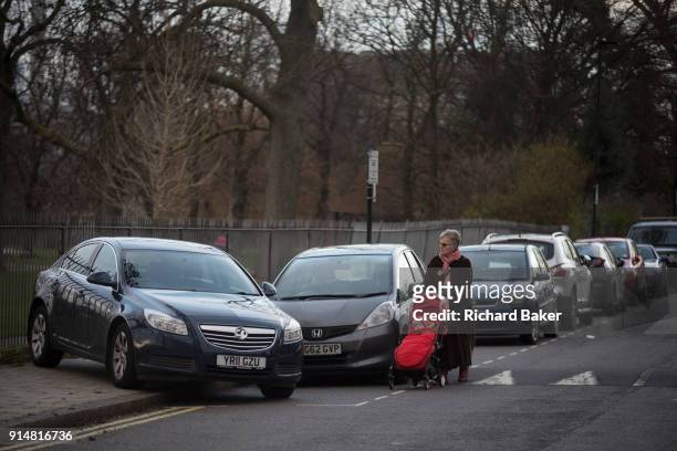 An elderly lady with a child's buggy walks in the road past a mysteriously abandoned Vauxhall car resting at 45 degrees, off the road but blocking a...