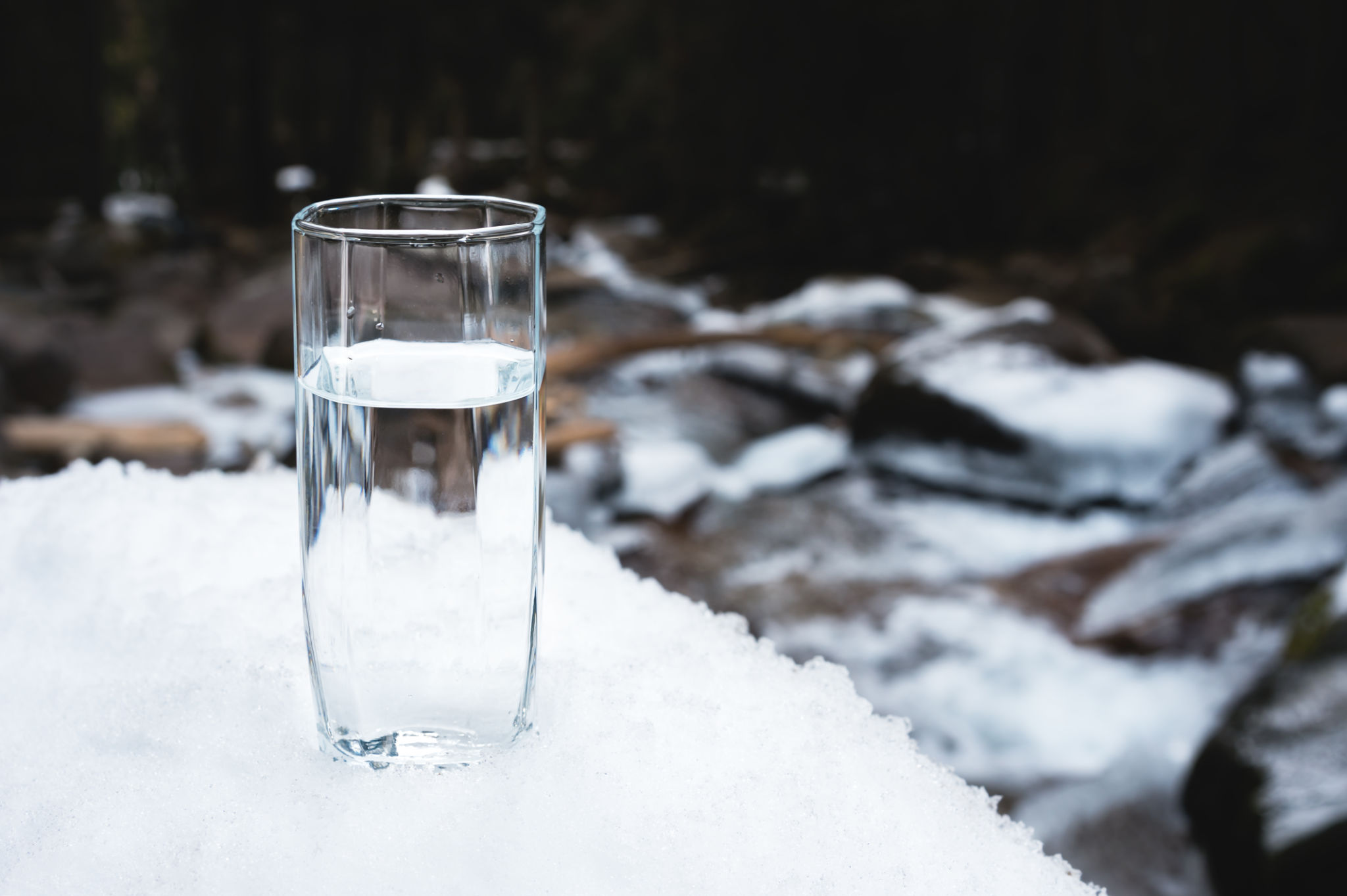 A transparent glass glass with drinking mountain water stands in the snow against a background of a clean mountain river and a forest in winter A transparent glass glass with drinking mountain water stands in the snow against a background of a clean mountain river and a forest in winter