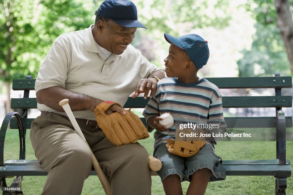 African grandfather and grandson holding baseball equipment on park bench