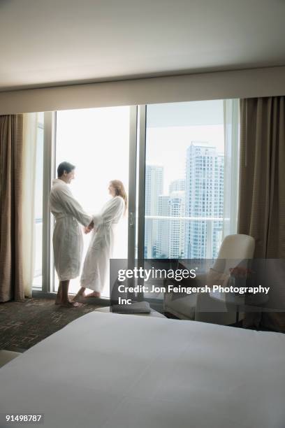 hispanic couple in bathrobes holding hands by window - confort at hotel bedroom stock pictures, royalty-free photos & images