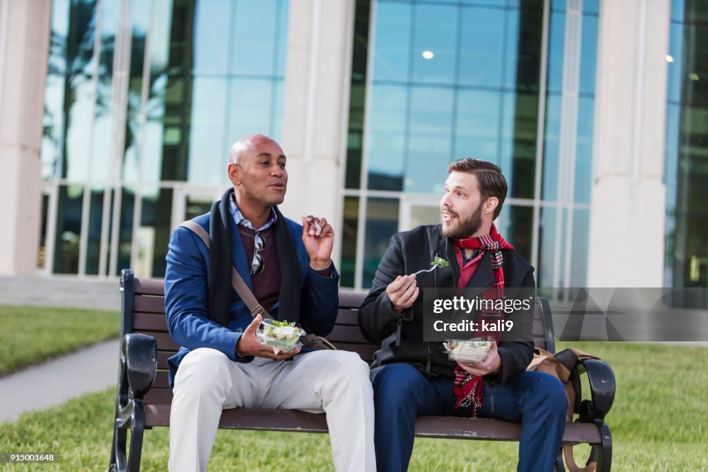 Two men eating lunch on bench outside office building