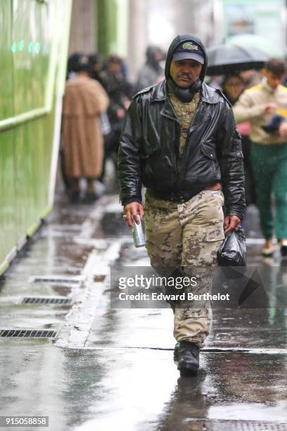 Guest wears a cap, a hood, a black blazer jacket, military camo print pants, under the rain, outside Paul Smith, during Paris Fashion Week - Menswear...