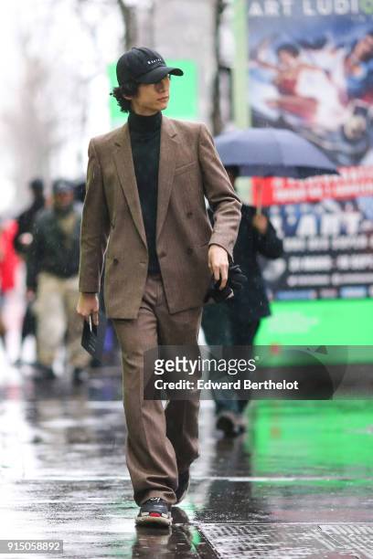 Guest wears a cap, a brown blazer jacket, suit pants, sneakers, a black turtleneck, under the rain, outside Paul Smith, during Paris Fashion Week -...