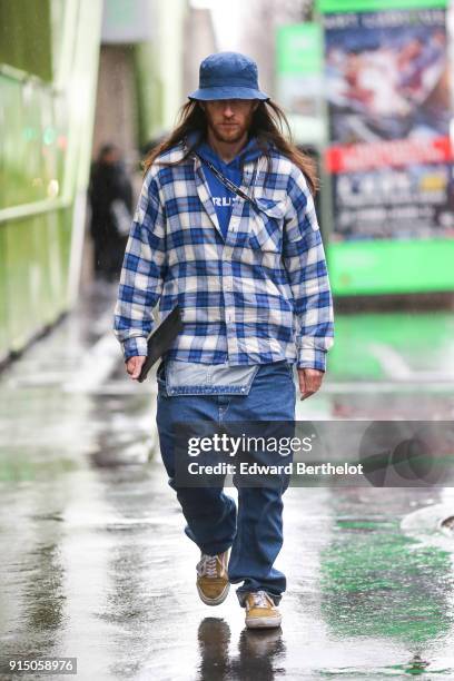 Guest wears a blue hat, a checked blue and white shirt, blue large denim jeans, yellow shoes, under the rain, outside Paul Smith, during Paris...