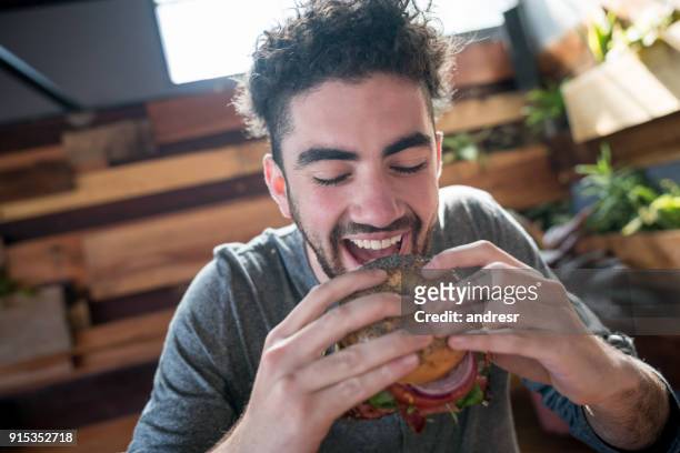 satiesfied young man taking a bite of a burger with eyes closed - bagels stock pictures, royalty-free photos & images