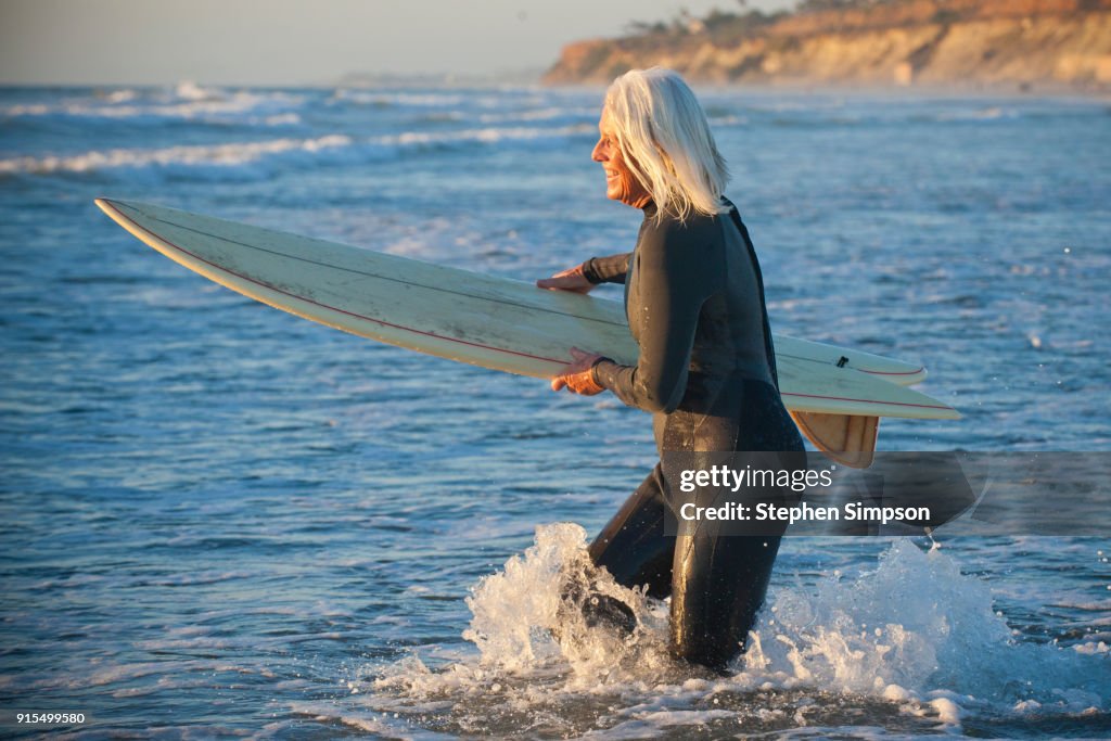 Fit gray-haired woman surfer in wetsuit running into the ocean