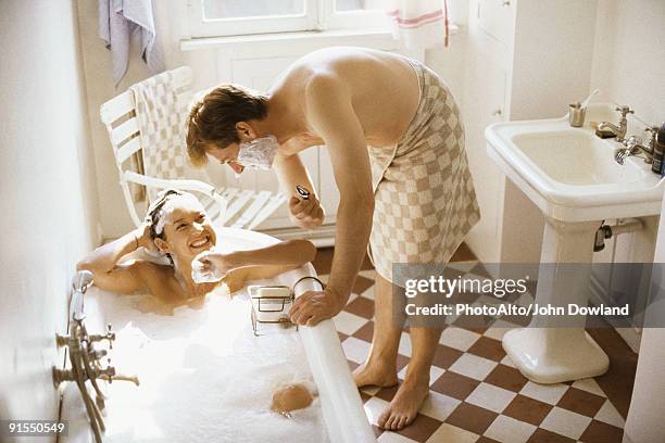 couple together in bathroom, woman in bathtub, man leaning over her with shaving cream on face - bad relationship stockfoto's en -beelden