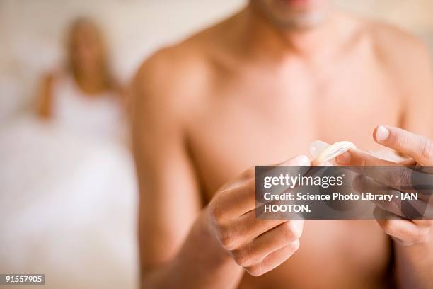 man unwrapping condom while girlfriend sitting in bed behind him - condoms stock pictures, royalty-free photos & images