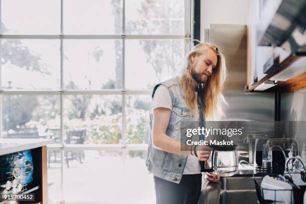 young professional making coffee at counter against window in brightly lit creative office - hipster in a kitchen stock-fotos und bilder