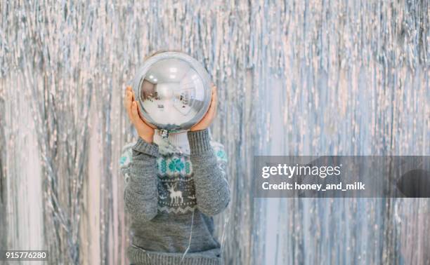 boy standing in front of silver tinsel decorated wall holding a giant christmas bauble - honey boy fotografías e imágenes de stock