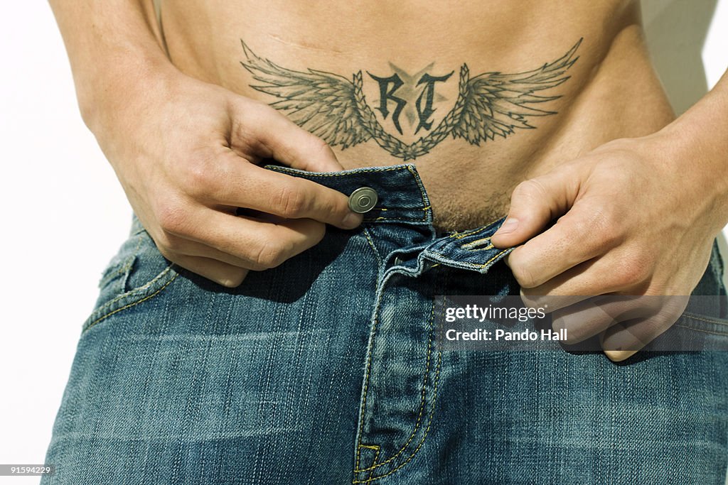 Young man with tattoo unbuttoning jeans, close-up