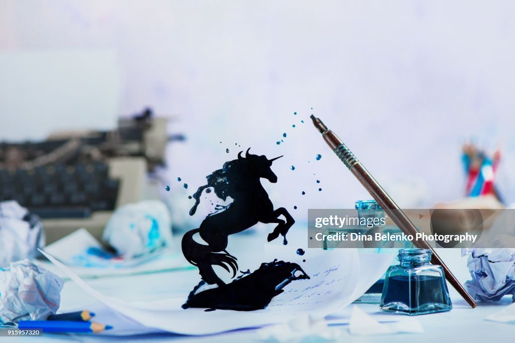 Fairytale writing prompt with unicorn silhouette made from splashes of ink from fountain pen and inkwell. Writer workplace with typewriter, pencils and papers on a light background with copy space. Creative writing concept with frozen motion.