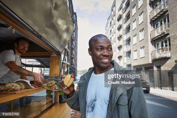 mid adult man having burger while standing at food truck in city - imbissstand stock-fotos und bilder