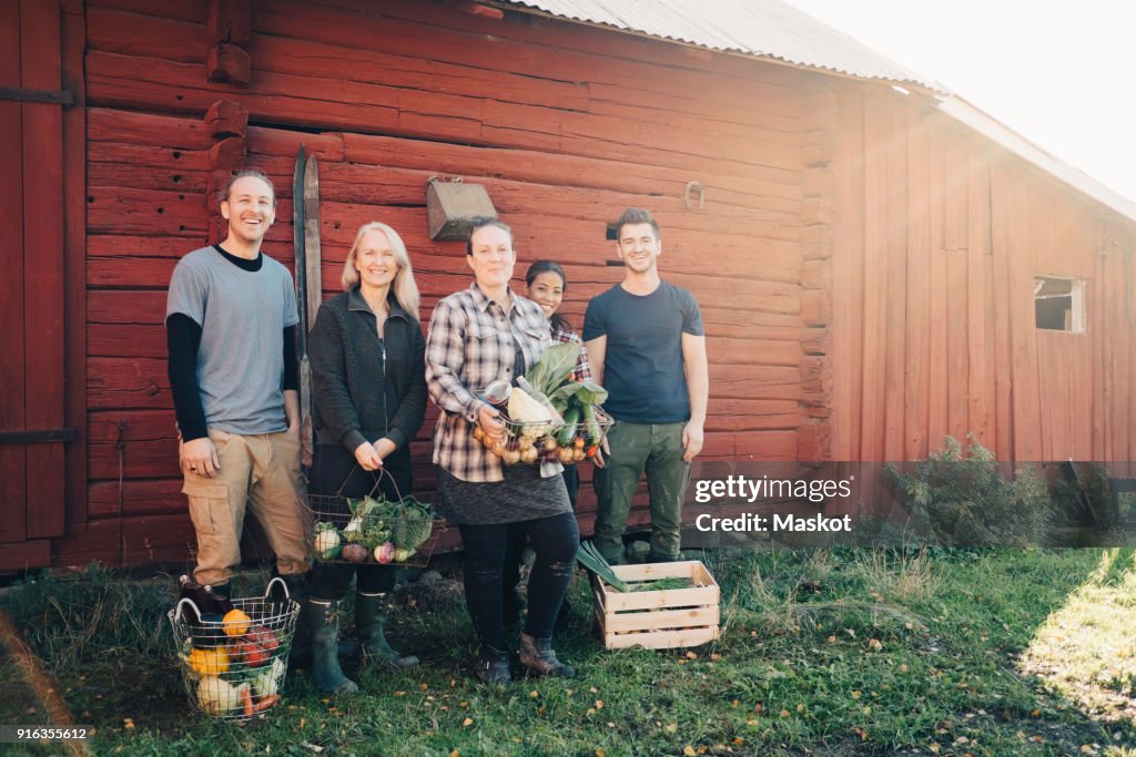 Full length of smiling farmers with organic vegetables standing against barn