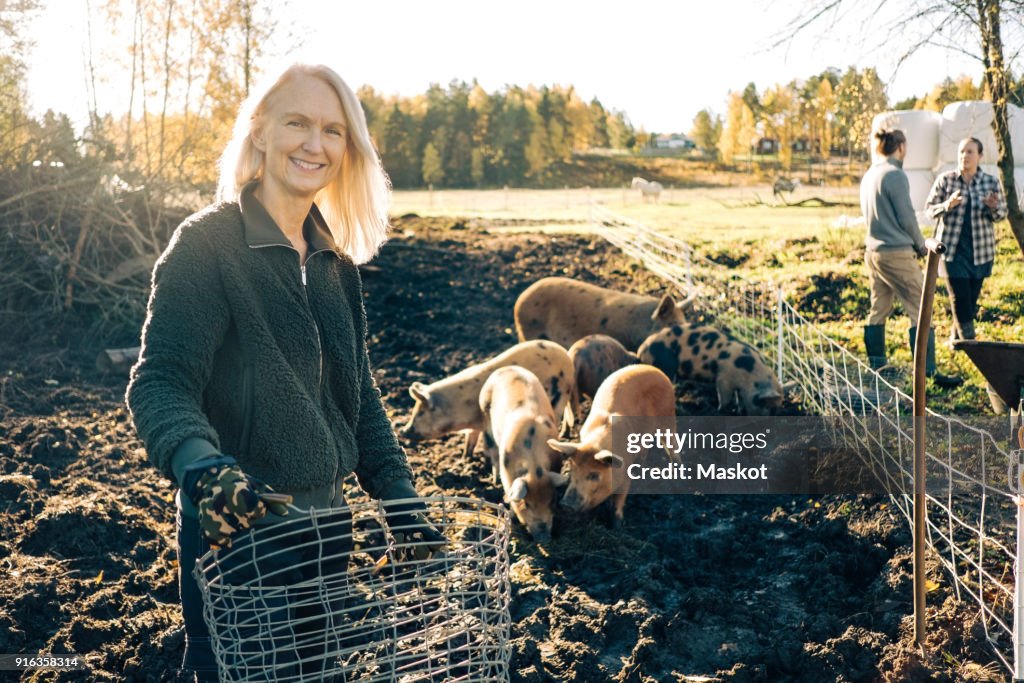 Portrait of smiling female farmer holding empty basket with pigs grazing at organic farm