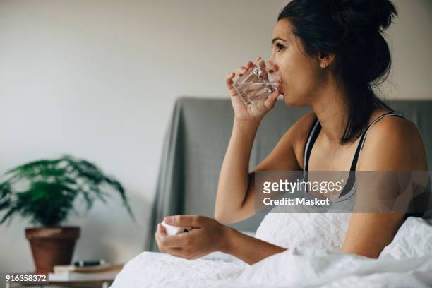 woman taking medicine while sitting on bed at home - drinking water glass woman stock-fotos und bilder