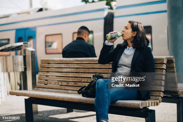 woman drinking juice while sitting on bench at platform by train - man woman train station stockfoto's en -beelden