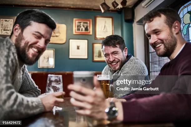 three british males sat together looking at a phone together laughing at it. - man sipping beer smiling photos et images de collection