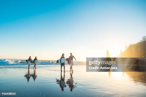 freunde steigen bei sonnenuntergang nach dem surfen aus dem wasser - surfer by the beach australia stock-fotos und bilder