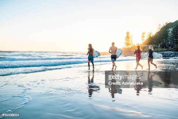 friends running into the water with surfboards at sunset - gold coast surfing stock pictures, royalty-free photos & images