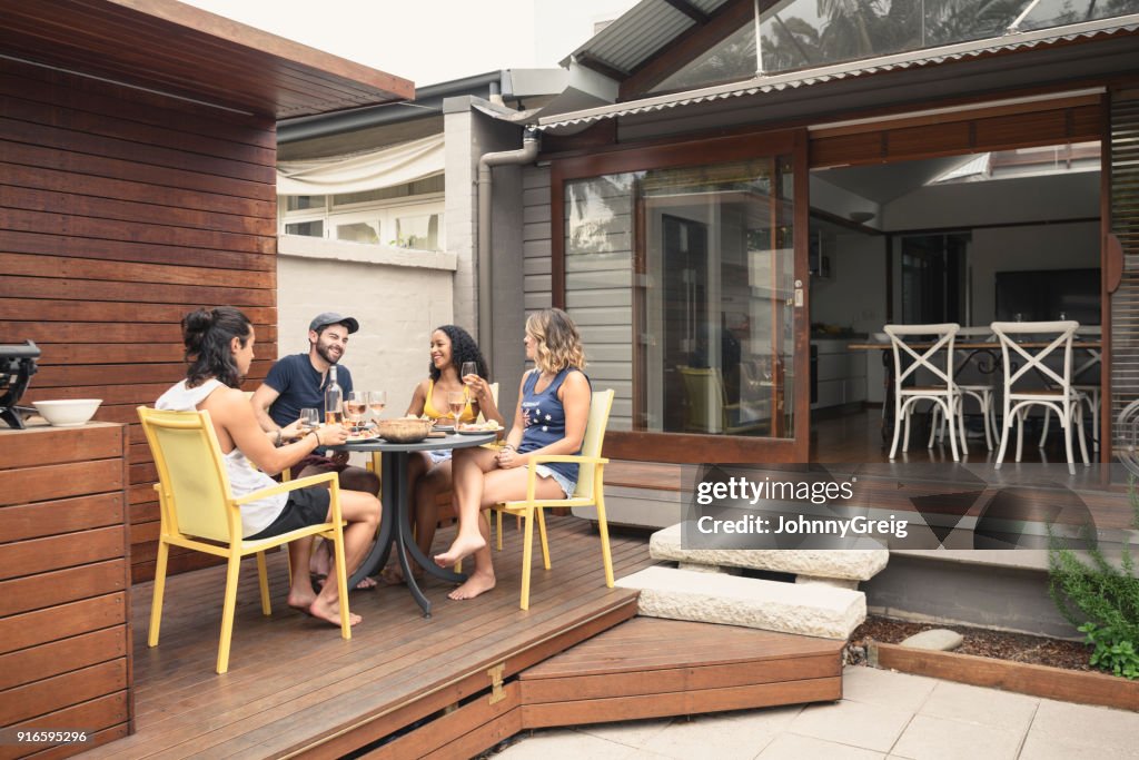 Group of four young adults relaxing on patio outside house with food and drink