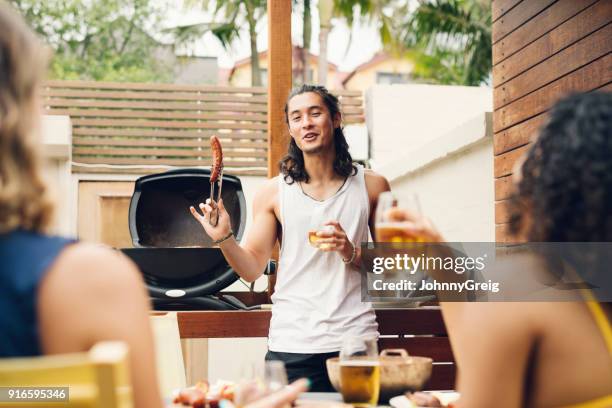 young man holding up sausage in front of barbecue with friends on foreground - up front party stock pictures, royalty-free photos & images