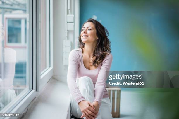 happy woman sitting beside window sill looking out of window - bien être photos et images de collection