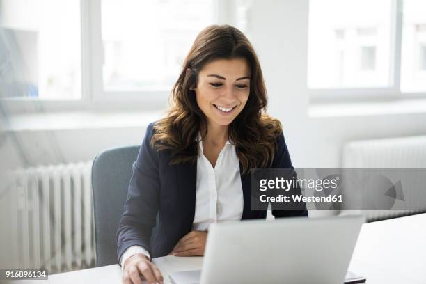 portrait of smiling businesswoman sitting at desk in the office working on laptop - capelli castani foto e immagini stock