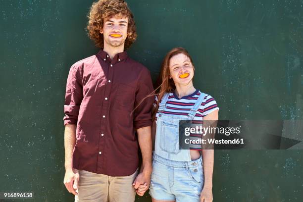 young couple standing in front of a green wall with orange slices in their mouth - freaky couples 個照片及圖片檔