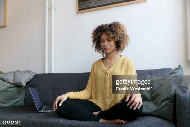 young woman sitting on couch at home next to laptop meditating - meditieren stock-fotos und bilder