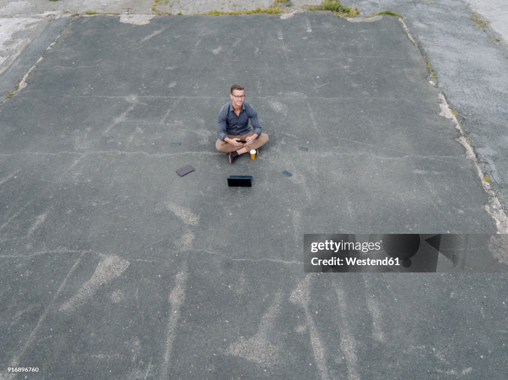 Young businessman with coffee to go and several mobile devices sitting on tarmac