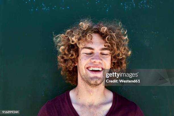 portrait of laughing young man with curly hair in front of a green wall - eyes closed stock-fotos und bilder