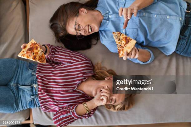 two laughing young women lying down eating pizza together - woman eating pizza stock-fotos und bilder