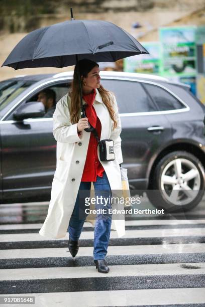 Guest wears a white trench coat, a red top, blue jeans, black leather shoes, holds an umbrella, under the rain, outside Paul Smith, during Paris...